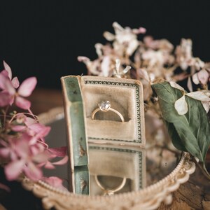 May include: A gold ring with a single diamond in a vintage ring box. The box is open and the ring is sitting inside. The box is surrounded by pink and white flowers.