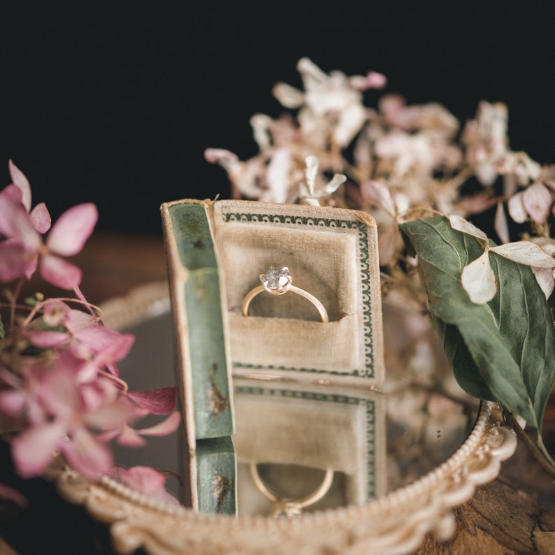 May include: A gold ring with a single diamond in a vintage ring box. The box is open and the ring is sitting inside. The box is surrounded by pink and white flowers.