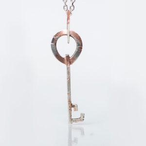 Puede incluir: Un collar con un colgante de llave de plata con un círculo de cobre en la parte superior. La llave tiene una superficie texturizada y un diseño simple.