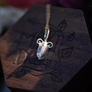 May include: A silver-colored pendant necklace featuring a small, textured amphora-shaped charm. The charm hangs from a delicate chain. The necklace is displayed on a dark wooden surface with the word "Design" visible.