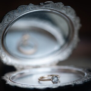 May include: A silver ring box with an ornate design, open to reveal a gold ring with a single diamond.