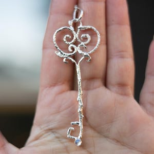 May include: A silver-colored key pendant with an ornate, heart-shaped top and a textured stem. The key features a small, round, iridescent stone at the bottom. The pendant is designed to be worn as jewelry.