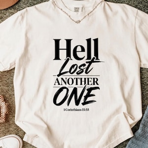 Peut inclure: Un t-shirt à manches courtes de couleur crème avec un texte noir indiquant Hell Lost ANOTHER ONE 1 Corinthians 15:55. Le t-shirt présente une encolure ronde et une coupe standard, avec le texte centré sur la poitrine dans des polices variées.