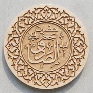 May include: A circular light-colored wooden plaque featuring intricate laser-cut patterns along the border. The center displays deeply engraved Arabic calligraphy, showcasing the name and title of Imam Jafar al-Sadiq in a clear, elegant script.