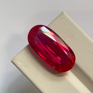 Puede incluir: Una gema de corte ovalado vibrante con un color rojo profundo y saturado. La piedra presenta múltiples facetas que crean reflejos nítidos y brillantes en su superficie pulida, mostrando un alto nivel de claridad y luminosidad.