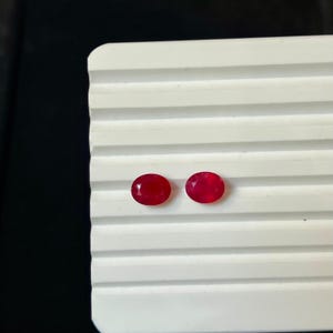 May include: Two oval-shaped, faceted red rubies with a polished finish. The gemstones are positioned side-by-side on a white, horizontally ribbed display surface. Each ruby exhibits a deep, vibrant red color with visible light reflection.
