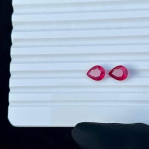Può includere: Due gemme sfaccettate a forma di pera, di un colore rosso intenso e vibrante, sono posizionate una accanto all'altra. Ogni pietra presenta una parte superiore appuntita e una base arrotondata, con una superficie lucida e sfaccettata.