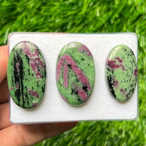 May include: Three oval-shaped cabochons of zoisite with ruby inclusions, featuring a vibrant green base color with distinct patches of deep magenta-red and streaks of black mineral deposits, arranged horizontally on a white foam display insert.