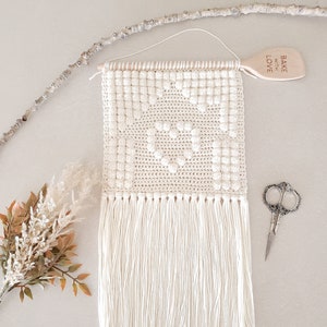 Op de afbeelding: Witte gehaakte wanddecoratie met een geometrisch ontwerp en franjes. De decoratie is bevestigd aan een houten stok. Een houten lepel met de tekst "Bake with Love" hangt aan de rechterkant van de afbeelding.