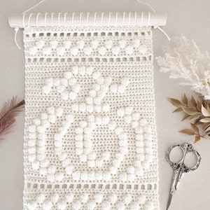 May include: A white crocheted wall hanging with a repeating pattern of raised circles. The hanging is attached to a wooden dowel rod.