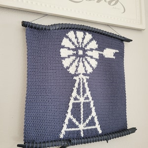 Op de afbeelding: Een gehaakte wandhanger met een wit windmolenontwerp op een donkerblauwe achtergrond. De hanger is ingelijst met twee donkerbruine takken.