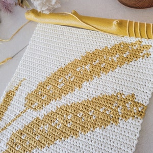 Op de afbeelding: Een wit en geel gehaakt wandkleed met een geometrisch patroon. Het wandkleed is gemaakt van dik garen en heeft bovenaan een natuurlijke houten lat.