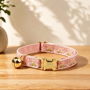 Può includere: Un collare per animali domestici rosa chiaro con un motivo floreale ricamato nei toni del rosa e del verde. Il collare include una fibbia in metallo dorato incisa con il nome Jinx e il numero 218-555-789, oltre a un piccolo campanello dorato.