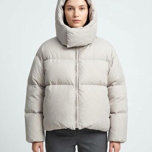 May include: A light gray puffer jacket featuring a high-neck hood with a front closure flap, long sleeves, and a quilted design. The jacket has a central zipper closure and is worn over dark gray trousers, providing a neutral and functional look.