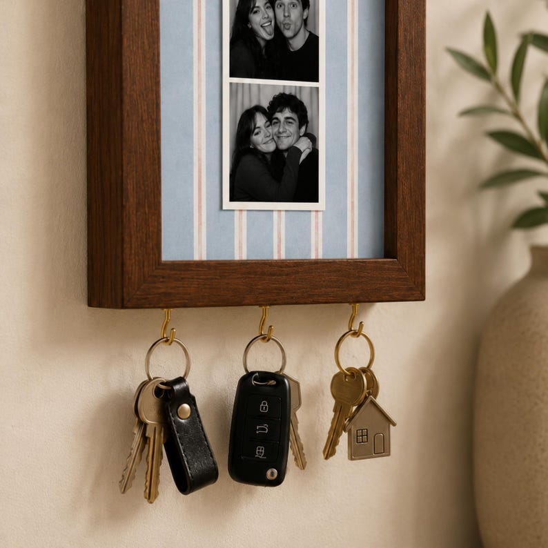 May include: A dark wood wall-mounted key holder featuring a vertical photo frame with a light blue and white striped background. The frame displays two black and white photo strips of a couple. Three gold hooks hold various keys and keychains.
