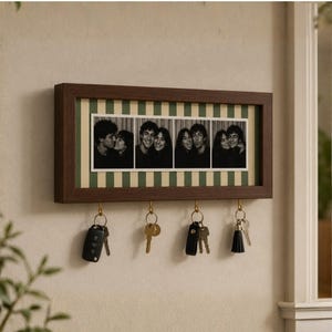 Pode incluir: Um porta-chaves de parede retangular em madeira escura com uma tira fotográfica horizontal de quatro retratos a preto e branco. A moldura tem um fundo às riscas verdes e creme e quatro ganchos de metal cor de latão com várias chaves.