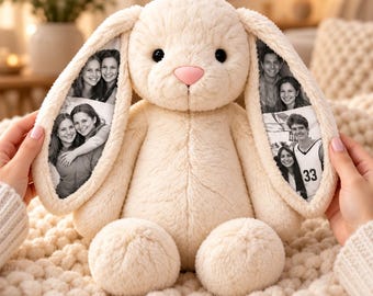 May include: A cream-colored plush rabbit toy with long, floppy ears. The inner side of each ear features printed black and white photographs of people. The rabbit has a soft, fuzzy texture, small black eyes, and a small, light pink nose.
