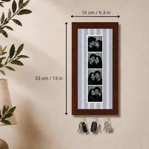 May include: A rectangular dark brown wooden wall-mounted key holder featuring a vertical photo strip of four black and white images. The frame measures 16 cm by 33 cm, or 6.3 inches by 13 inches. It includes three metal hooks for hanging keys.