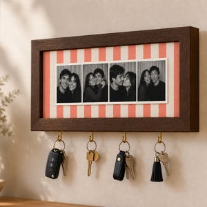 May include: A rectangular dark brown wooden wall-mounted key holder featuring a central horizontal display area with a coral and white striped background. Four gold-toned metal hooks hang below, each holding a set of keys with various keychains.