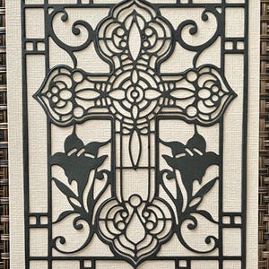 May include: A black, intricately cut cross design with floral accents and a decorative border. The cross is centered on a cream-colored background, creating a striking contrast. The design is mounted on a textured, woven surface.