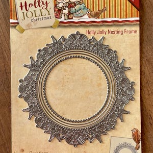 May include: A silver-colored Holly Jolly Nesting Frame cutting die. The die features a circular design with intricate snowflake and floral patterns. The packaging includes illustrations of children and a bird, with the text "Holly Jolly Christmas."