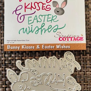 May include: A metal die set for crafting with the words "Bunny Kisses & Easter Wishes" in a cursive font. The set includes a bunny graphic and the words "bunny kisses & easter wishes". The die set is approximately 3.6" x 2.4".