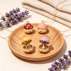 Peut inclure: Quatre colliers médaillons en forme de champignon sur des chaînes dorées. Deux sont fermés, avec des chapeaux rouge et rose à pois blancs. Deux sont ouverts, révélant une abeille sous un chapeau jaune et une sorcière sous un chapeau violet.