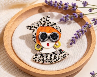 May include: A decorative brooch shaped like a stylized head wearing a black and white checkered headscarf, round black sunglasses with glittery frames, and yellow hoop earrings. The figure has bright orange hair, a white face, and red lips.