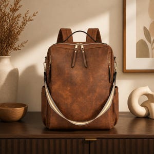 Puede incluir: Una mochila marrón con acabado liso y mate, que cuenta con un asa superior, dos correas para los hombros y una correa cruzada desmontable con un patrón de rayas blancas, amarillas y grises. Incluye cremalleras doradas y bolsillos laterales.