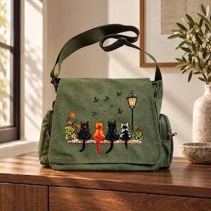 May include: An olive green canvas messenger bag featuring a printed graphic of four cats sitting on a ledge next to a lit street lamp. The cats are black, orange, black, and black and white. Small birds are flying above the cats near the lamp.