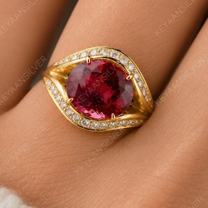 May include: A gold ring featuring a large, round, deep red gemstone at the center, surrounded by a halo of small, clear, sparkling stones. The band has a curved, split-shank design that tapers toward the sides, set with additional small stones.
