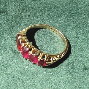 Puede incluir: Un anillo de oro con cinco piedras preciosas rojas de corte ovalado dispuestas en fila. La banda de metal presenta detalles intrincados de volutas en los laterales e incluye grabados de sellos visibles en la superficie interior del anillo.