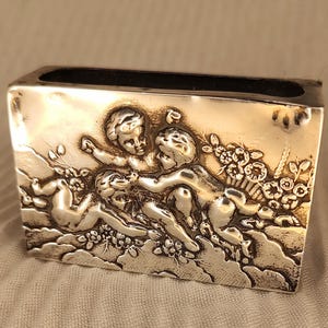 May include: A rectangular silver-toned metal matchbox cover featuring a detailed embossed relief of three cherubs playing among clouds and floral elements. The metallic surface has a polished finish with dark recessed areas highlighting the figures.