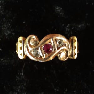 Könnte beinhalten: Ein goldener Ring mit einem zentralen, rund geschliffenen roten Rubin, der von zwei kleineren, runden, klaren Steinen flankiert wird. Die Ringschiene zeigt ein geschwungenes Design mit texturierten Metallakzenten und vertikalen Rillen.