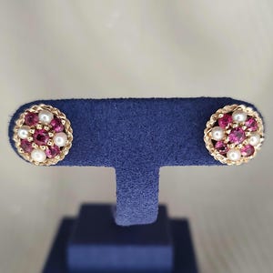 May include: A pair of round stud earrings featuring a central deep pink gemstone surrounded by a cluster of smaller white pearls and additional pink gemstones, all set within a textured gold-toned metal frame with a scalloped edge design.