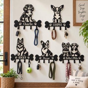 May include: Five black metal wall-mounted key and leash holders shaped like dog bones, each featuring a laser-cut dog silhouette and a personalized name. The holders include multiple hooks for hanging keys, leashes, and accessories, with various pet items attached to the hooks.