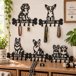 Könnte beinhalten: Fünf schwarze, wandmontierte Leinen- und Schlüsselhalter in Knochenform, jeweils mit einer Hundesilhouette und einem Namen: Charlie, Cooper, Rocky, Oscar und Bella & Milo. Jeder Halter verfügt über vier Haken zum Aufhängen von Gegenständen.