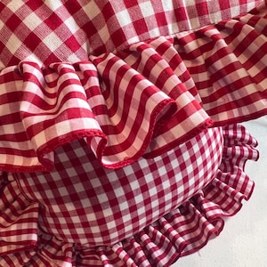 May include: A decorative pillow with a red and white gingham pattern. The pillow features a ruffled edge in the same fabric. The fabric is a classic check design, creating a cozy and country-style aesthetic.