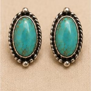 Peut inclure: Une paire de boucles d'oreilles à clous ovales avec des pierres de turquoise polies présentant des motifs de matrice brune. Chaque pierre est sertie dans une monture en métal argenté avec une bordure torsadée et des perles sphériques.