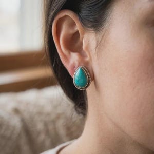 Peut inclure: Une boucle d'oreille en forme de goutte avec une pierre turquoise sertie dans un cadre en argent doté d'une bordure en corde torsadée. La pierre présente des veines sombres naturelles sur sa surface bleu-vert et une finition argentée.