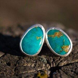 Peut inclure: Une paire de boucles d'oreilles à clous présentant des pierres de turquoise de forme ovale avec des motifs de matrice brune naturelle. Chaque pierre est enchâssée dans un serti clos en métal argenté lisse et poli pour un rendu épuré.