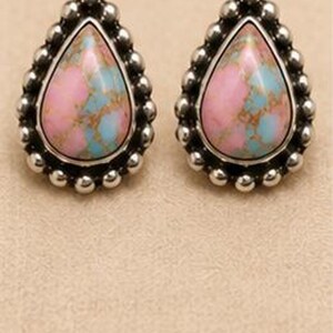 Peut inclure: Une paire de boucles d'oreilles à clous en forme de goutte, dotées d'une pierre centrale au motif marbré rose et bleu clair, entourée d'une bordure décorative composée de petites sphères en métal argenté disposées en cadre perlé.