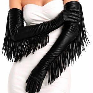May include: A pair of long black opera-length gloves reaching above the elbows. The gloves feature a smooth finish and are adorned with long, thin black fringe detailing that runs along the outer edge from the wrist up to the upper arm area.