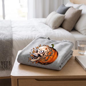May include: A light grey crewneck sweatshirt featuring a graphic print of a human skull emerging from a carved orange pumpkin. The skull has wide, startled eyes and an open mouth, with orange pumpkin shards floating around the central design.