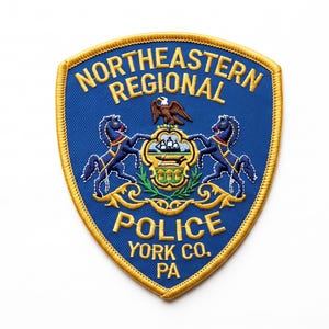 May include: A shield-shaped embroidered patch with a blue background and gold border. It features the text NORTHEASTERN REGIONAL POLICE YORK CO. PA, an eagle, and two horses flanking a central emblem containing a ship and a plow on a green field.
