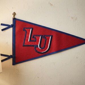 May include: A triangular felt pennant in red with a blue border and two blue ties on the left side. The center features the letters LU embroidered in red with white and blue outlines. The pennant is displayed hanging from a small metal hook.
