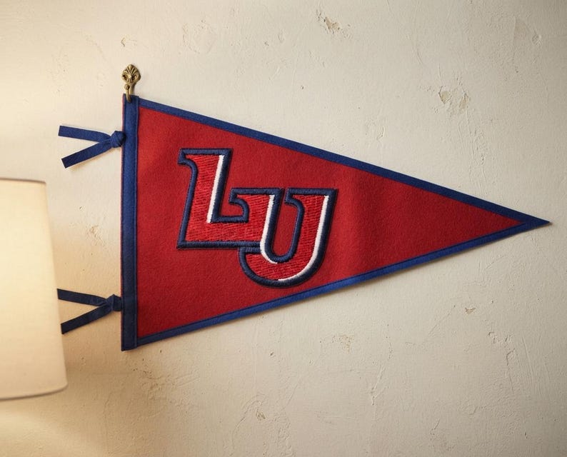 May include: A triangular felt pennant in red with a blue border and two blue ties on the left side. The center features the letters LU embroidered in red with white and blue outlines. The pennant is displayed hanging from a small metal hook.
