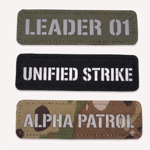 Puede incluir: Tres parches rectangulares de tela con bordes cosidos. El parche superior es de color verde oliva con el texto LEADER 01. El parche central es negro con el texto UNIFIED STRIKE. El parche inferior presenta un patrón de camuflaje con el texto ALPHA PATROL.