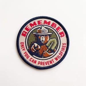 May include: A circular embroidered patch featuring Smokey Bear wearing a hat with the text SMOKEY. The bear holds a shovel and points forward. The border text reads REMEMBER ONLY YOU CAN PREVENT WILDFIRES in red lettering on a beige background.