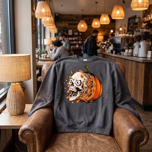 May include: A charcoal grey crewneck sweatshirt featuring a graphic print of a white human skull emerging from a bright orange pumpkin. The skull has wide, startled eyes and an open mouth, with orange pumpkin fragments floating around the design.